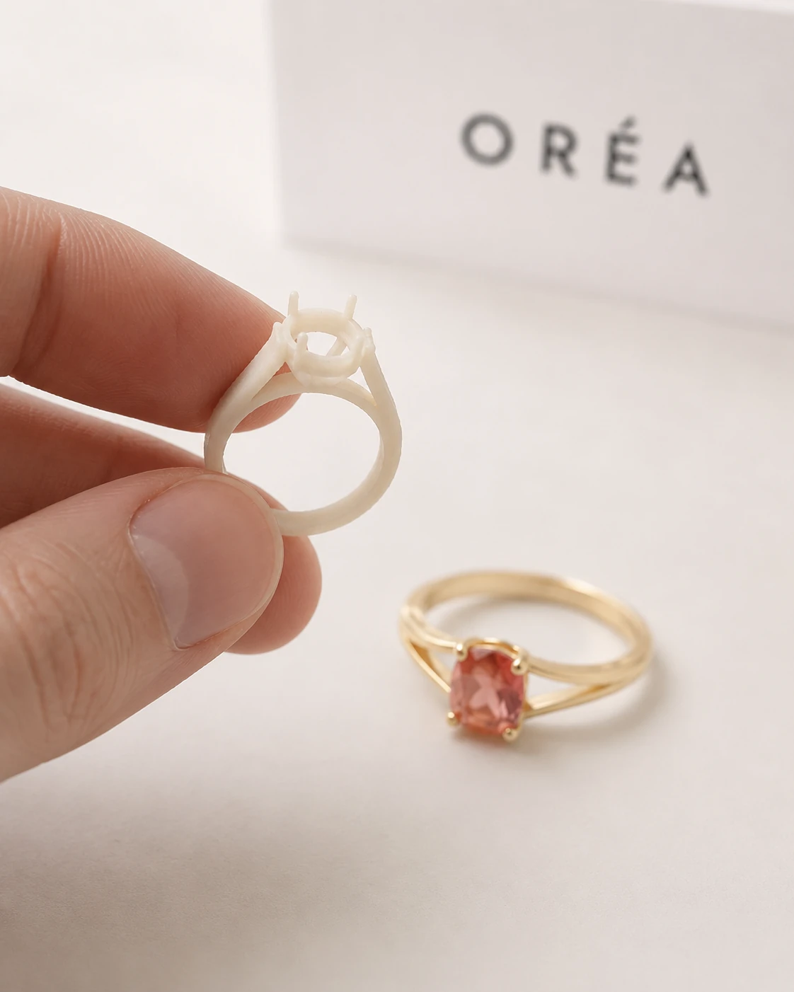 A bone white resin proof of the ring held between finger and thumb, its claws cut but unset, the finished gold ring set with the orange pink sapphire resting on the sweep behind
