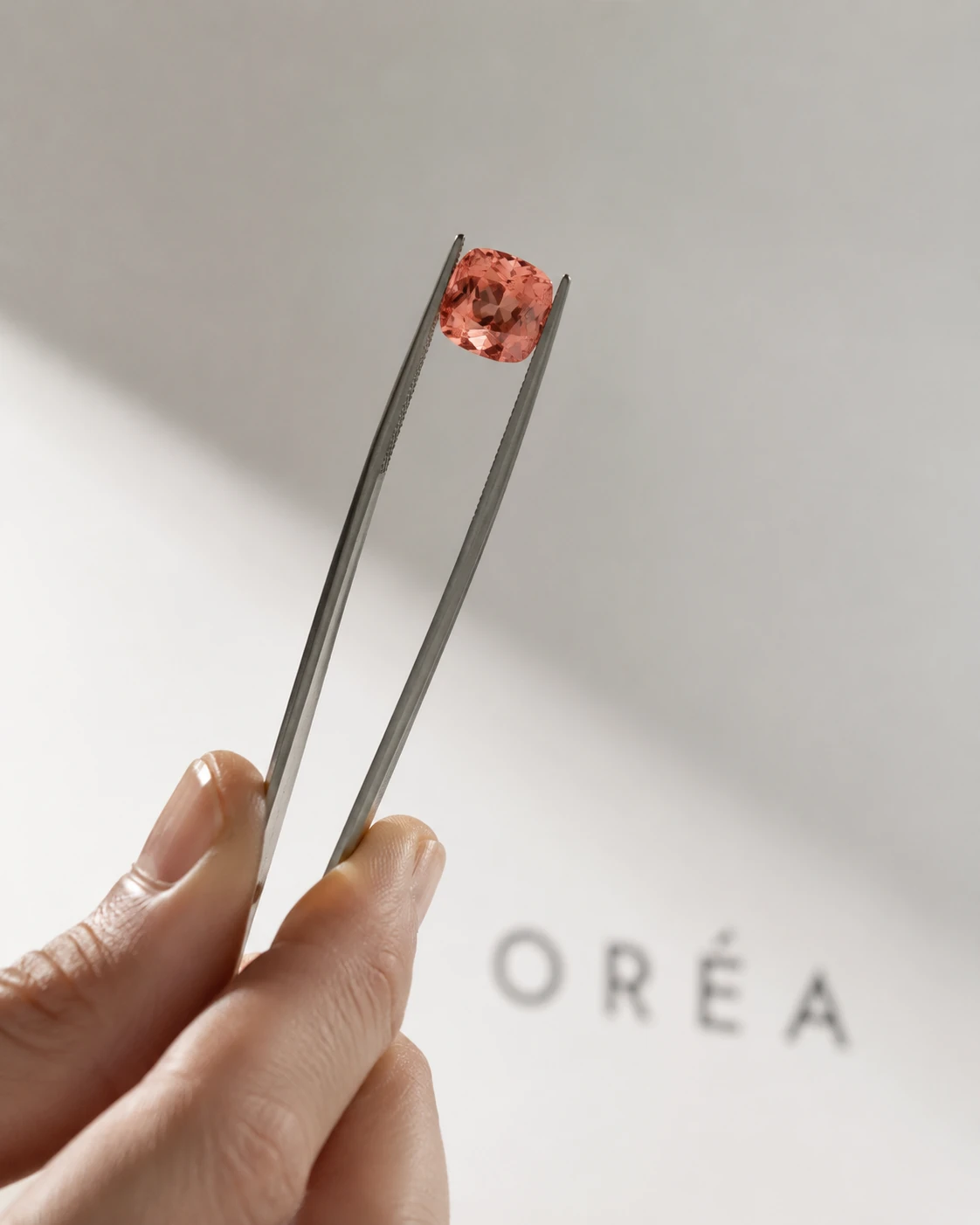 The same orange pink cushion cut sapphire held close in tweezers, lit through so the colour glows