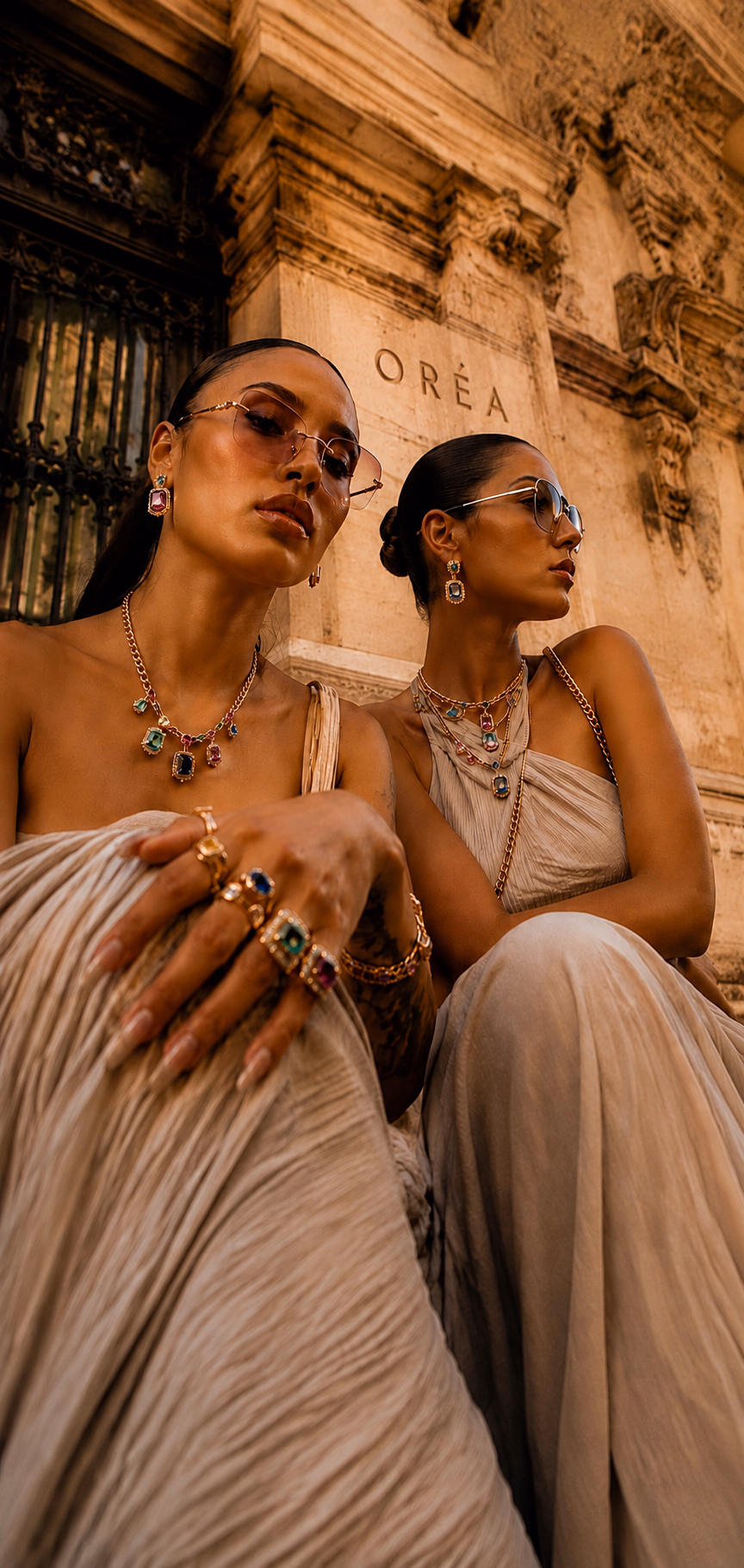 Two women in pleated stone-coloured dresses and tinted glasses wearing gem-set gold necklaces, drop earrings and stacked rings, seated before a carved façade